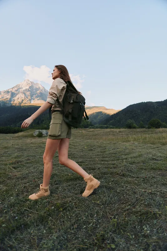 Woman with backpack hiking in an open meadow near mountains, casual outdoor wear. Wide landscape, sense of adventure, wellness, and exploration. Soft natural light creates a cheerful mood.