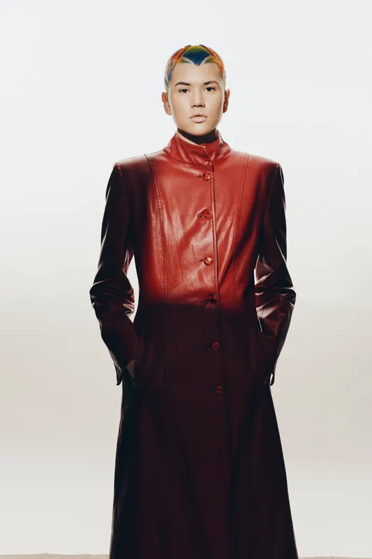 young woman with colorful short hair wearing a long burgundy leather coat, standing confident with hands in pockets against plain white background