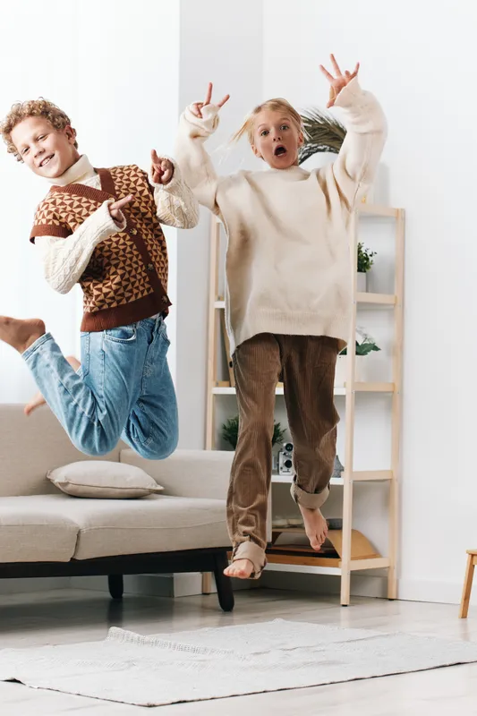 Two playful children jump mid air indoors, showcasing carefree energy and candid happiness as they wear cozy sweaters and jeans in a bright living room setting.