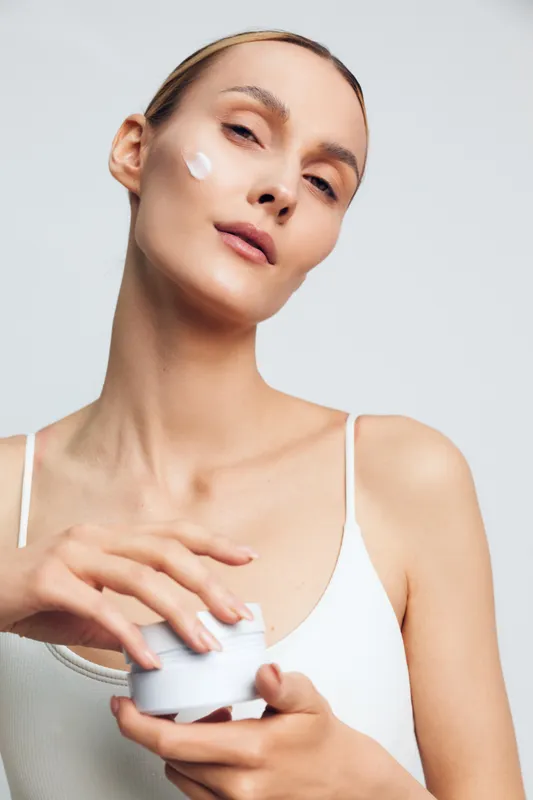 Young woman applying facial cream on cheek, skincare routine, clean skin, natural beauty, soft lighting, white background, casual outfit, healthy lifestyle concept