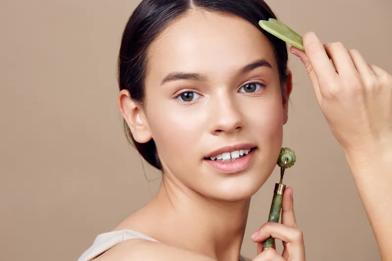 Young woman with smooth skin using a jade roller for facial massage, showcasing natural beauty and skincare routine, against a neutral beige background
