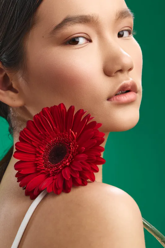 asian woman portrait with a vivid red gerbera flower near her shoulder, set against a green backdrop, expressing elegance, calm confidence, and modern beauty.