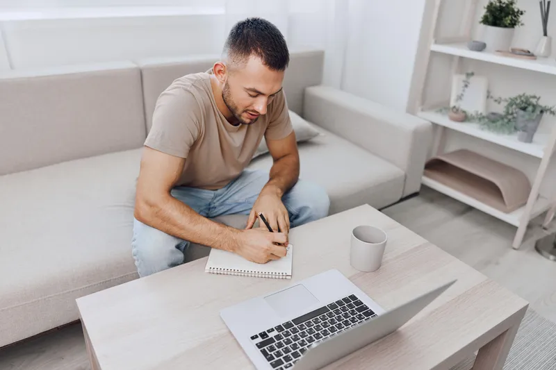 Focused man writing in a notebook while sitting at a modern workspace. Soft neutral colors enhance the cozy atmosphere of productivity and creativity.