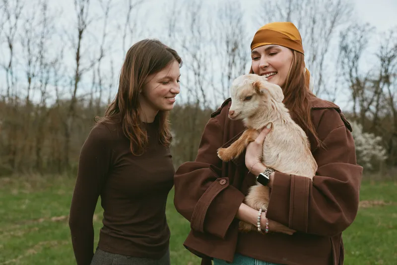 Two women outdoors share a warm moment with a goat kid, embracing nature and friendship, casual fashion and rustic charm in a sunny field.