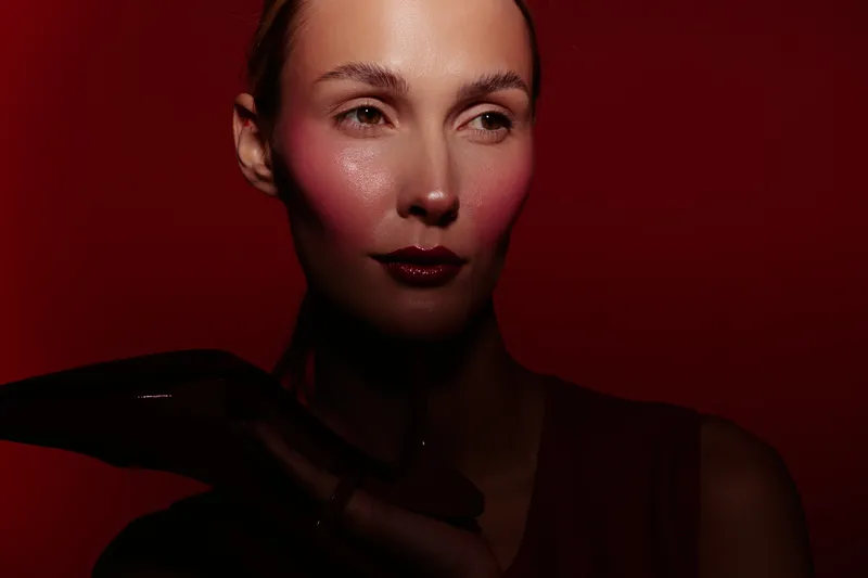 Portrait of a woman with glossy lips and glowing skin illuminated by red light. Dark background and dramatic shadows create a mysterious and artistic mood in the studio.
