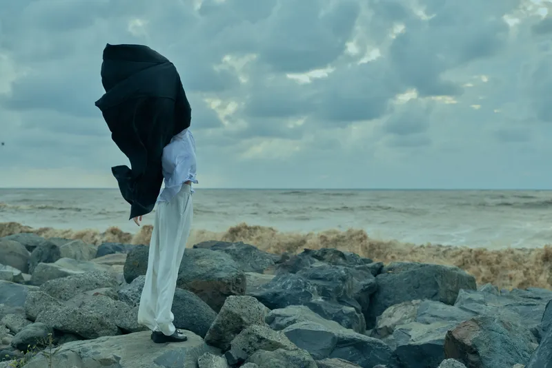 Windswept figure on a rocky shore, cloak billowing toward the sea, creating a dramatic moody scene with stormy clouds and a sense of isolation