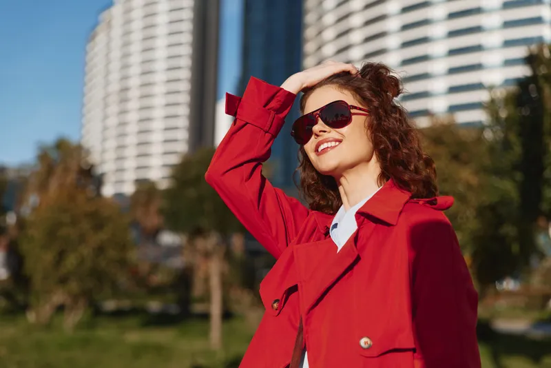 Happy woman in red coat and sunglasses enjoys sunny day outdoors smiling with hand on head near modern buildings in urban park setting.