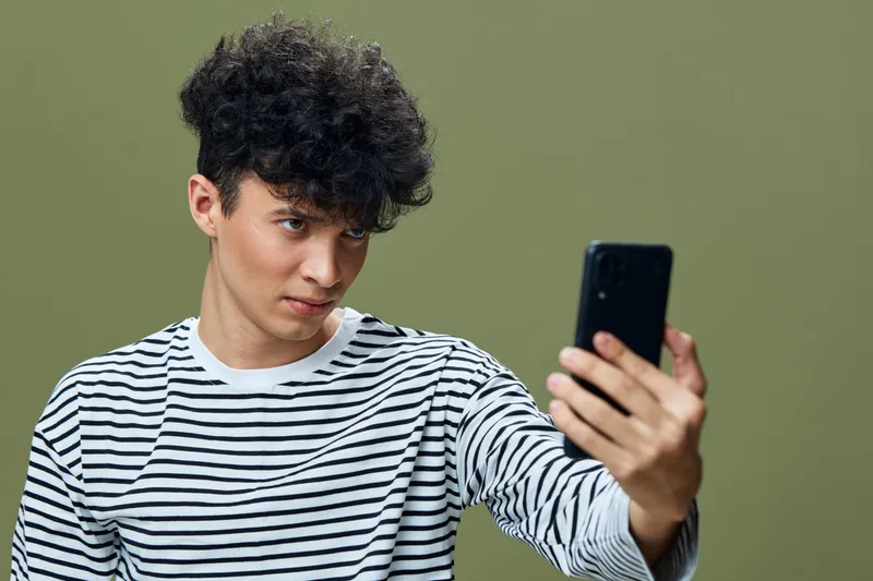 young man with curly hair wearing striped shirt taking selfie with smartphone, green background, casual style, focused expression, modern technology use