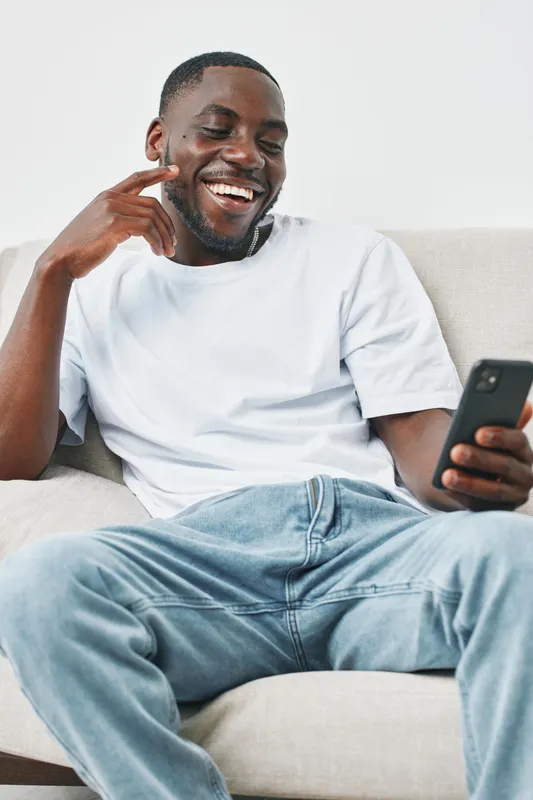 Smiling young African American man relaxed on a sofa, looking at his smartphone, wearing casual white T shirt and jeans, conveying joy and comfort in a minimalist setting