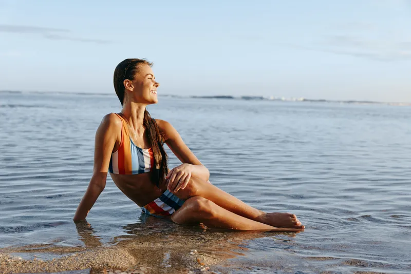 Woman sitting in shallow ocean water on beach shoreline, colorful swimwear, smiling in summer daylight, calm sea waves, visible horizon, outdoor vacation relaxation, serene mood