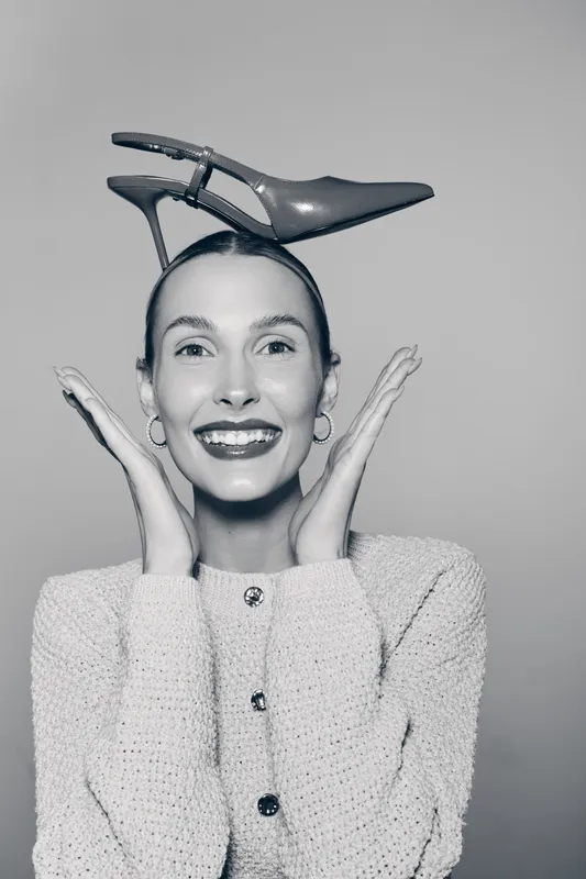 Happy Caucasian woman with light skin smiling and holding a high heel shoe on her head. Concept of fashion, joy, and playful mood in studio.