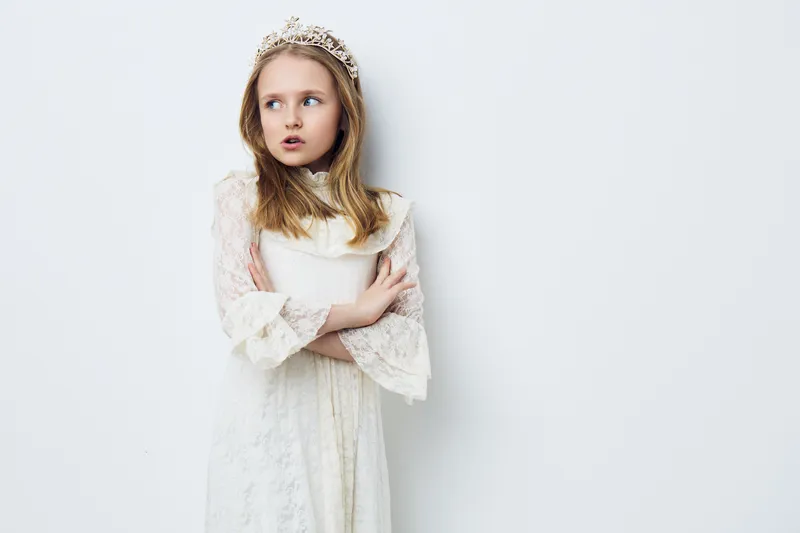 Portrait of a young girl in a vintage white dress with a tiara, looking thoughtful against a light background, embodying innocence and curiosity