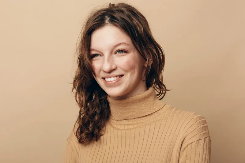 Smiling woman with wavy brown hair wearing beige turtleneck sweater looks away on beige background. Concept of natural beauty, casual style, and positive mood in studio portrait.