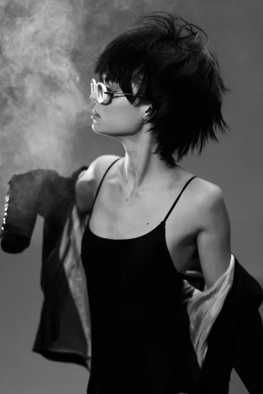 Black and white portrait of a stylish woman with short dark hair holding a smoking object, wearing glasses and a sleeveless dress.