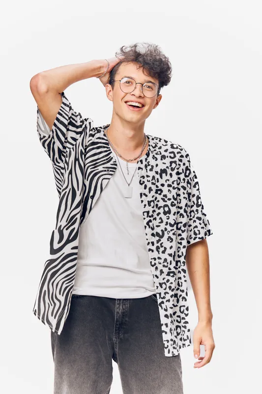 young man smiling in stylish black and white animal print shirt with glasses and curly hair, casual modern look on white background, joyful and relaxed