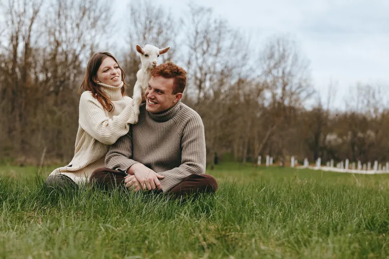 Young couple in cozy sweaters sits on green grass with a playful baby goat on the man's shoulders enjoying a relaxed outdoor lifestyle in early spring.