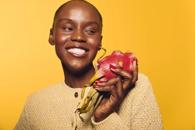 Smiling black woman with dark skin holding a red dragon fruit against yellow background. Concept of healthy lifestyle, natural food, and positive emotions in studio lighting.
