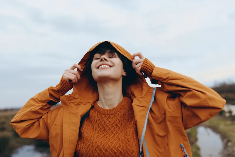 Smiling woman lifestyle portrait outdoors, adjusting hood of orange jacket in autumn wind by a river, knit sweater visible, soft clouds in sky, calm everyday warmth natural light