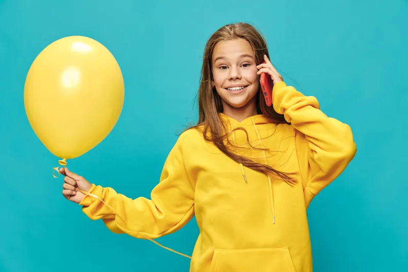 Girl wearing yellow hoodie holding yellow balloon and talking on phone against blue background smiling. Child, communication and cheerful with long hair in casual clothes.