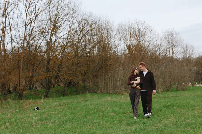 Couple walking outdoors with a kid goat nearby, sharing affection in a grassy park. A warm lifestyle moment in nature, with smiling faces, gentle breeze, and a close family connection.