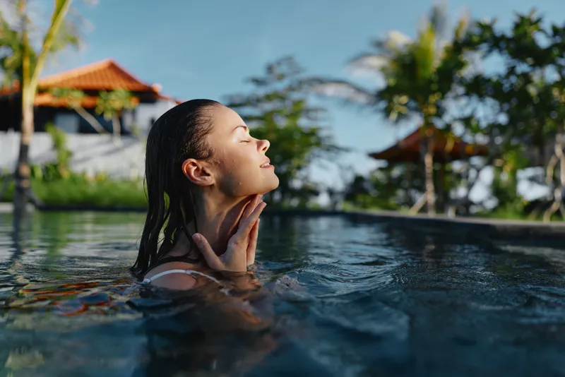 Vibrant woman in a sunlit pool, eyes closed, enjoying relaxation outdoors, tropical backdrop, clear water, lush greenery, warm breeze, casual summer mood and carefree happiness