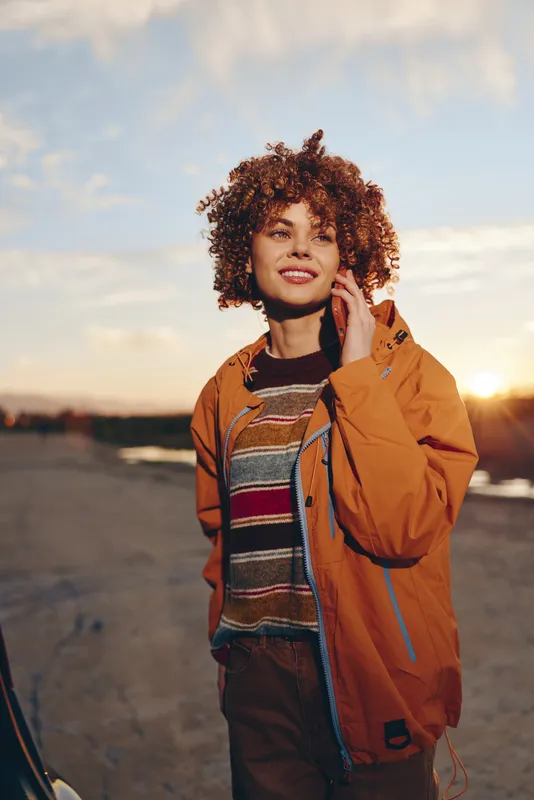 Woman lifestyle outdoors at sunset, portrait of a smiling young woman with curly hair, wearing an orange jacket and casual fashion, standing on a road with a soft sky, warm sunlight