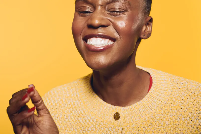 Close-up of a happy black woman with dark skin smiling with closed eyes against yellow background. She wears a yellow sweater and shows positive emotions and joyful mood.