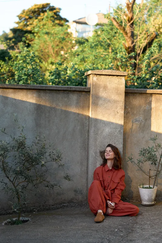 A young woman in a red jumpsuit sits on a concrete terrace beside a weathered wall, gazing upward with a reflective, peaceful mood and quiet composure