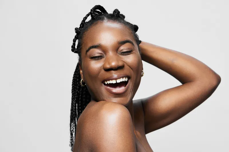 Joyful young woman with braided hair smiling brightly against a light background, exuding happiness and positivity in a minimalist fashion