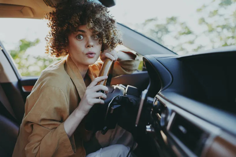 Young woman with curly hair sits in the driver's seat, gripping the steering wheel with a surprised, focused expression inside a modern car, ready for a spontaneous road trip