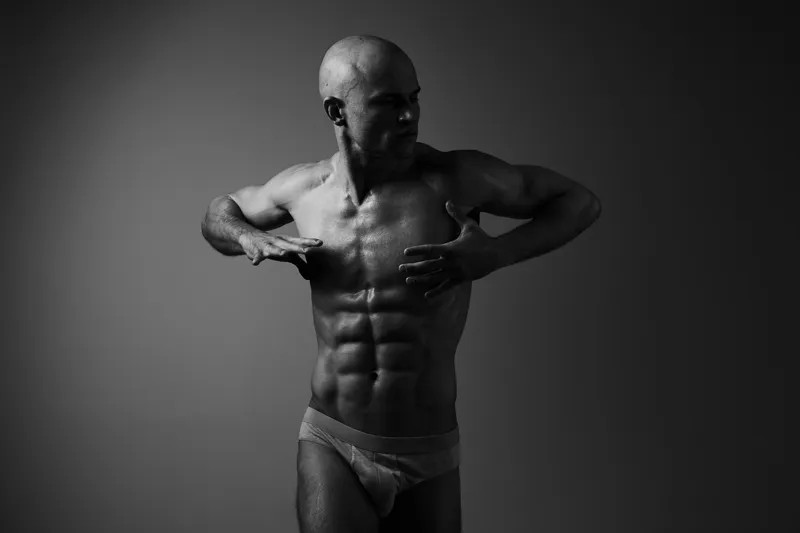 Black and white image of a muscular man posing confidently, showcasing his physique and strength with dramatic lighting.
