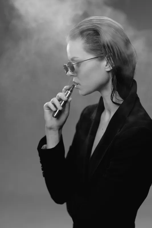Black and white portrait of a stylish woman vaping with sunglasses and smoke.