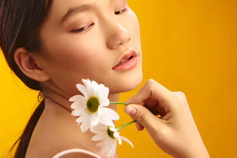 Asian woman gracefully holds white daisies against a bright yellow backdrop, exuding calm, beauty, and a soft studio mood with gentle lighting and graceful pose.