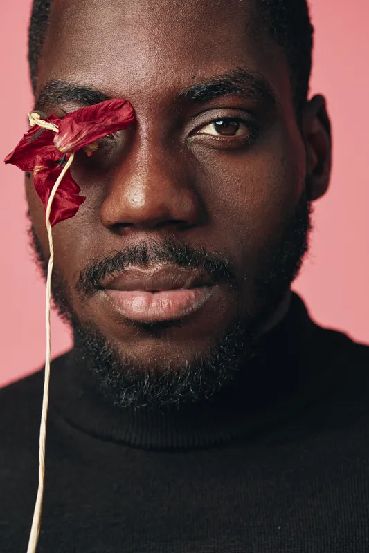 African man with dark skin and beard holding red dried flower near eye in creative conceptual visual style. Closeup portrait with pink background and black turtleneck sweater for artistic expression.