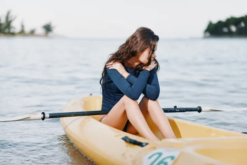 Young woman sits in a yellow kayak on calm water, hugging her knees and looking downward, creating a peaceful, contemplative moment on the lake.