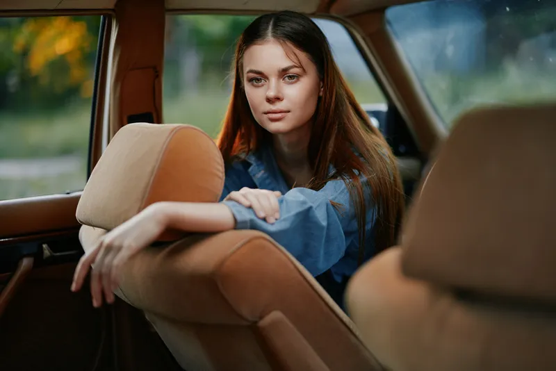 Young woman with long hair in a blue shirt, sitting in a vintage car, looking contemplative and serene, surrounded by a soft, natural background.