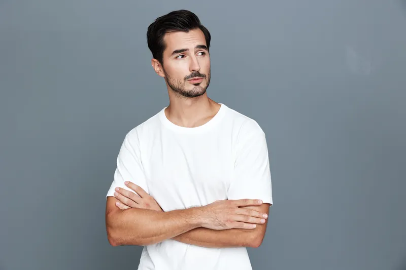 Thoughtful young caucasian man with stylish haircut wears white t-shirt standing against gray background, arms crossed, showcasing a contemplative expression.