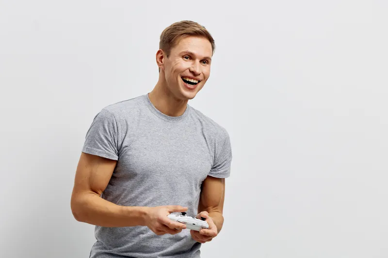 Joyful smiling young man holding a game controller, casual grey t-shirt, cheerful expression, enjoying gaming activity, isolated on plain white background, studio portrait, lifestyle concept, happines