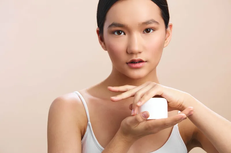 asian woman applies skincare in a clean studio, smiling softly as she holds a small jar and looks ahead, conveying calm beauty routine and confident self care.