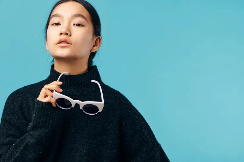 Young woman in black sweater holding white sunglasses against blue background. Portrait of stylish female model with natural makeup and confident expression in casual outfit.