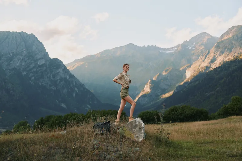 Young woman hiking in mountains stands on rock in open field with tall grass under cloudy sky during daytime, enjoying active lifestyle and nature outdoors.