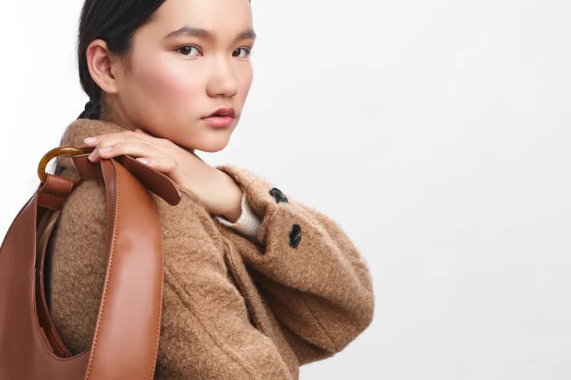 asian woman wearing a cozy wool coat and a leather bag over her shoulder, exudes calm confidence and chic style in a clean studio setting.