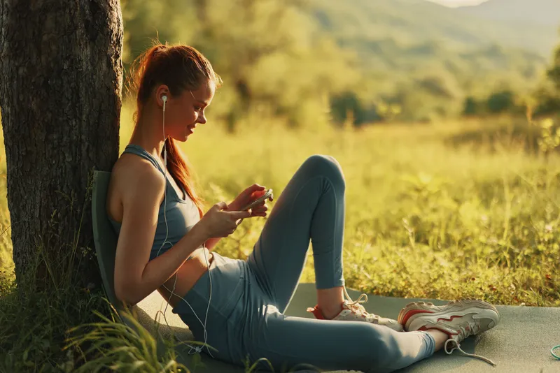 Young athletic woman enjoying music outdoors in nature, wearing casual fitness attire, representing a healthy lifestyle and positive emotions in a vibrant green landscape