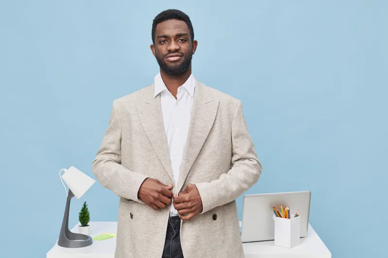 Confident young man with beard and curly hair dressed in beige blazer and white shirt standing in bright modern office with blue background, holding hands together, professional business look, workspa