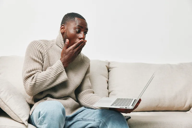 A surprised young Black man in a cozy sweater sitting on a sofa, using a laptop, expressing disbelief or shock, showcasing emotions and casual lifestyle