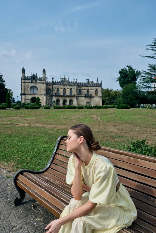 Woman in a pale yellow dress sits thoughtfully on a wooden bench in a sunny park with a grand castle in the background, creating a calm outdoor scene.