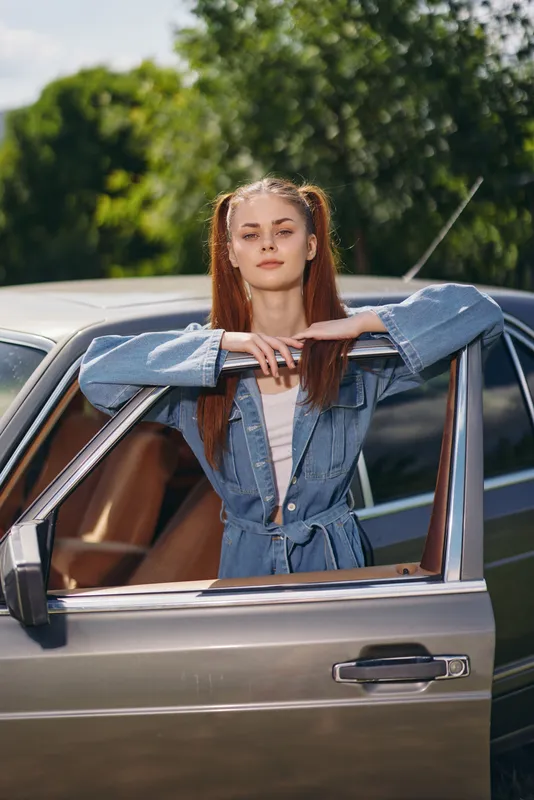 Young woman with long red hair in denim jacket poses by vintage car, exuding confidence and style against a lush green backdrop, embodying summer vibes.