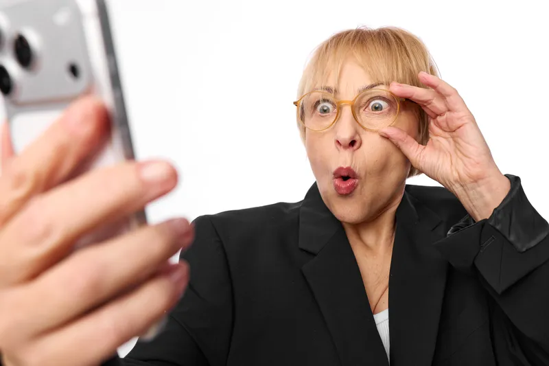 Surprised business woman in office with glasses, hand on head, open mouth reaction, blurred coworker with smartphone, concept of shock, communication, anxiety and stress