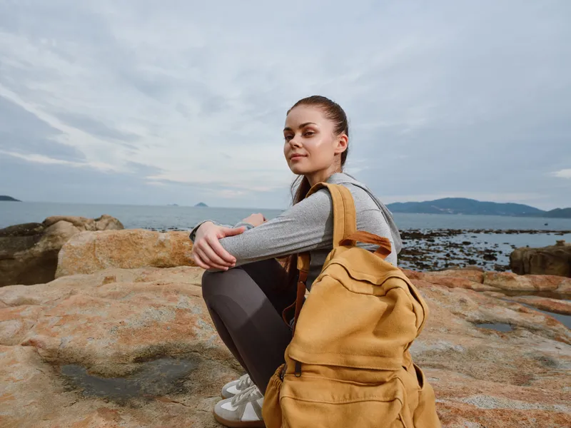 Young woman with a yellow backpack sitting on rocks by the sea, showcasing a relaxed and contemplative mood, perfect for travel or adventure themes