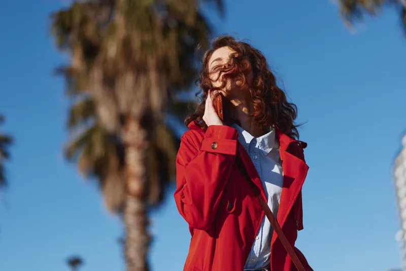 A young woman in a red coat enjoys a sunny day outdoors with her hair blowing in the wind, standing near a palm tree under a clear blue sky.